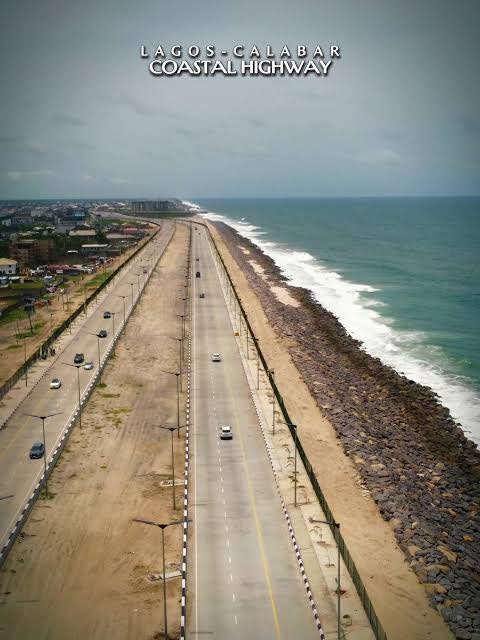 Aerial view of the completed Lagos–Calabar Coastal Highway running beside the Atlantic, with rock armour protecting the seaward edge and built-up areas inland.