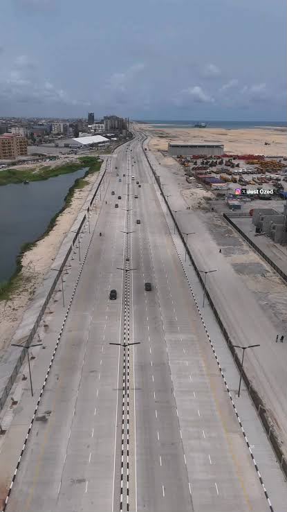 Aerial view along a wide multi-lane section of the Coastal Highway in traffic, with a lagoon inlet on one side and industrial yards on the other.